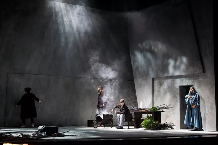 L'Annonce faite à Marie Un personnage fait du feu au centre d'une pièce aux murs gris. De la fumée s'élève. À droite, une femme en bleu tient un bébé. Près du feu, un homme reste debout, regardant à gauche. À gauche, un personnage n'est vu que de dos.