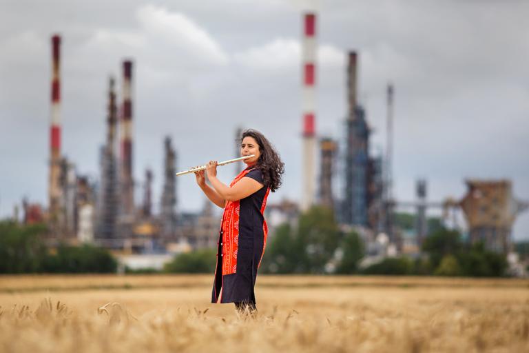 Naïssam Jalal Portrait de Naïssam Jalal jouant de la flûte traversière dans un champ de blé, une usine en arrière plan